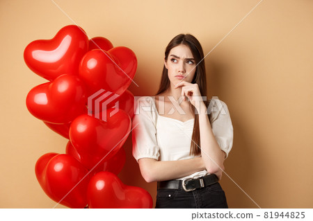 Valentines day and love concept. Thoughtful young woman standing near hearts balloons and looking aside with doubtful face, having assumptions, beige background Valentines day and love concept. Thoughtful young woman standing near hearts balloons and looking aside with doubtful face, having assumptions, beige background 81944825