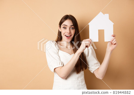 Real estate. Excited woman got new apartment, showing paper house cutout and smiling happy, standing on beige background Real estate. Excited woman got new apartment, showing paper house cutout and smiling happy, standing on beige background 81944882