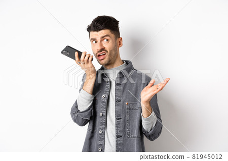 Stylish handsome man talking on speakerphone, record voice message with smartphone near lips, discuss something and looking aside at empty space, standing on white background Stylish handsome man talking on speakerphone, record voice message with smartphone near lips, discuss something and looking aside at empty space, standing on white background 81945012