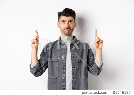 Sad frowning guy pointing and looking up, standing hesitant on white background Sad frowning guy pointing and looking up, standing hesitant on white background 81946123