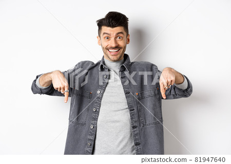 Excited smiling male model pointing fingers down, showing logo with happy face, demonstrate cool promo, standing on white background Excited smiling male model pointing fingers down, showing logo with happy face, demonstrate cool promo, standing on white background 81947640