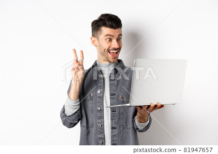 Funny young man video chat on laptop, showing v-sign and smiling at computer camera, standing against white background 81947657