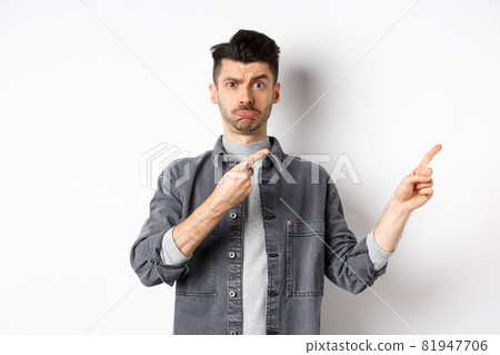 Sad caucasian guy pointing fingers right and pouting, looking hesitant, standing on white background 81947706