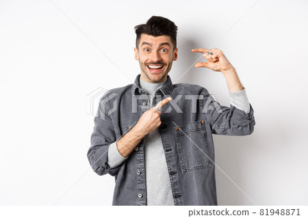Excited man showing small size, pointing finger at little thing and smiling, standing against white background Excited man showing small size, pointing finger at little thing and smiling, standing against white background 81948871