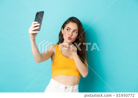 Technology and lifestyle concept. Stylish beautiful woman posing for selfie, sending air kiss at camera, using photo filters app on mobile phone, standing against blue background Technology and lifestyle concept. Stylish beautiful woman posing for selfie, sending air kiss at camera, using photo filters app on mobile phone, standing against blue background 81950772