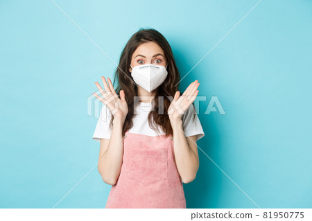 Covid-19, vaccination and quarantine concept. Excited and surprised young woman in medical respirator, face mask from coronavirus, clap hands and look amazed at camera, blue background 81950775