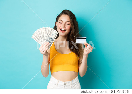Good-looking smiling girl holding credit card, looking pleased and determined at dollar bills money, standing against blue background Good-looking smiling girl holding credit card, looking pleased and determined at dollar bills money, standing against blue background 81951906