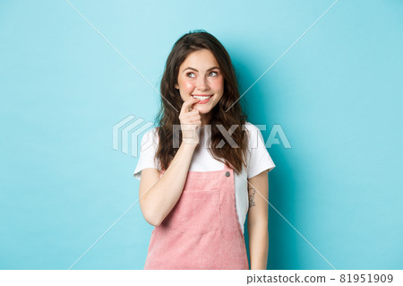 Thoughtful silly girl with cute curly hairstyle, biting finger and looking left at promo logo, smiling thinking about tasting ot trying something, standing over blue background Thoughtful silly girl with cute curly hairstyle, biting finger and looking left at promo logo, smiling thinking about tasting ot trying something, standing over blue background 81951909