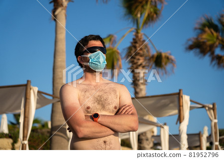 Man in medical mask stands alone on empty beach near sun loungers and palm trees on Cyprus, Paphos city during coronavirus quarantine and lockdown. New normal in vacation time after COVID-19 pandemic 81952796