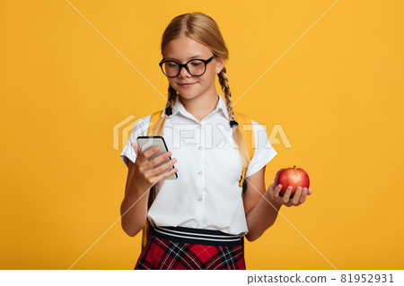 Happy young blonde girl pupil with pigtails and backpack in glasses typing on phone, holds red apple 81952931
