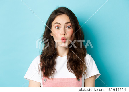 Close up portrait of excited stylish girl with curly hairstyle and beauty make up, pucker lips intrigued and stare at camera with surprised face, standing over blue background 81954324
