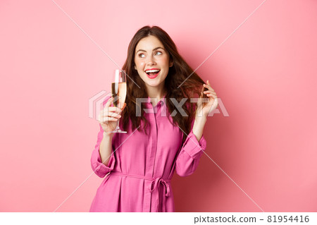 Celebration and holidays concept. Stylish coquettish woman in trendy dress, holding glass of champagne at party, smiling pleased, standing over pink background Celebration and holidays concept. Stylish coquettish woman in trendy dress, holding glass of champagne at party, smiling pleased, standing over pink background 81954416