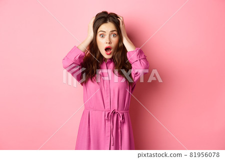 Shocked glamour girl gasping and looking startled, holding hands on head, standing in panic against pink background 81956078