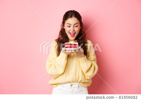 Celebration and holidays. Happy birthday girl stares at delicious bday cake dessert and smiles, stands against pink background Celebration and holidays. Happy birthday girl stares at delicious bday cake dessert and smiles, stands against pink background 81956082