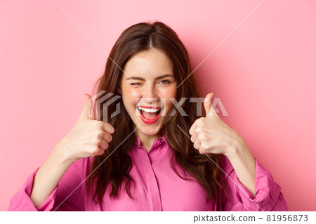 Cheeky glamour girl winking at you and shows thumbs up, praise great work, good choice, standing over pink background 81956873