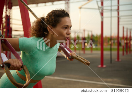 Close-up of young athletic woman with perfect body in sportswear doing push-ups with fitness suspension straps in the sportsground. Attractive African American woman working out outdoors at sunrise. 81959075