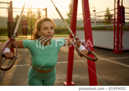 Close-up portrait of attractive determinate mixed race woman training arms with stationary fitness straps outdoors, doing push-ups, training upper body, chest, shoulders, pectoral muscles, triceps 81959082