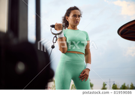 Attractive determinate Latin American female athlete performing weight training, doing exercises on biceps, flexing arms on a cable crossover gym machine outdoor Attractive determinate Latin American female athlete performing weight training, doing exercises on biceps, flexing arms on a cable crossover gym machine outdoor 81959086