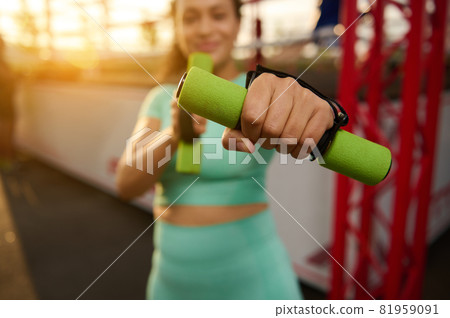 Soft focus on sporty woman's outstretched hand holding a small dumbbell, standing on the background of a boxing arena. Blurred fitness woman exercises outdoors at sunrise 81959091