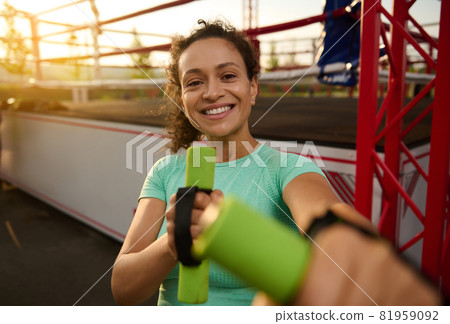 Portrait of beautiful Latin American attractive young sporty woman smiling with toothy smile, enjoying the morning cardio workout at sunrise, posing with dumbbells on the background of a boxing arena 81959092
