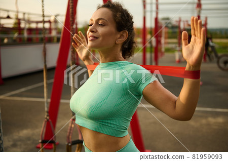 Beautiful slender sporty girl doing training with rubber resistance band on a sportsground. Attractive African American, mixed race fit woman stretches her back and shoulders muscles. 81959093