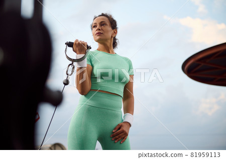 Attractive determinate Hispanic female athlete performing weight training, doing exercises on biceps, flexing arms on a cable crossover gym machine outdoor Attractive determinate Hispanic female athlete performing weight training, doing exercises on biceps, flexing arms on a cable crossover gym machine outdoor 81959113