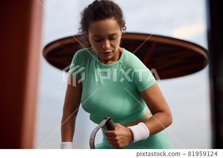 Bodybuilding concept. Sporty determinate young Hispanic female athlete performing a weight training with stationary cable crossover equipment in the outdoor sport field 81959124