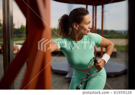 Beautiful and attractive woman of Latin American ethnicity doing exercise on triceps and shoulders muscles with stationary outdoor cable crossover. Determinate athlete working out in sportsground Beautiful and attractive woman of Latin American ethnicity doing exercise on triceps and shoulders muscles with stationary outdoor cable crossover. Determinate athlete working out in sportsground 81959126
