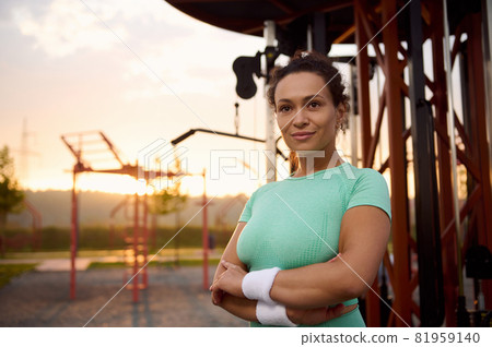 Close-up portrait of attractive mixed race, gorgeous Hispanic sportswoman, female athlete in sportswear standing with crossed arms on the background of outdoor gym machines in the morning at sunrise. Close-up portrait of attractive mixed race, gorgeous Hispanic sportswoman, female athlete in sportswear standing with crossed arms on the background of outdoor gym machines in the morning at sunrise. 81959140