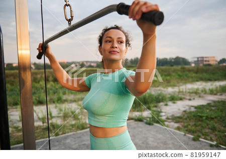 A smiling slender Latin American athlete woman flexing muscles on gym machine. Sporty woman executing exercise with crossover exercise-machine in outdoor sports field. Fitness, bodybuilding concept 81959147