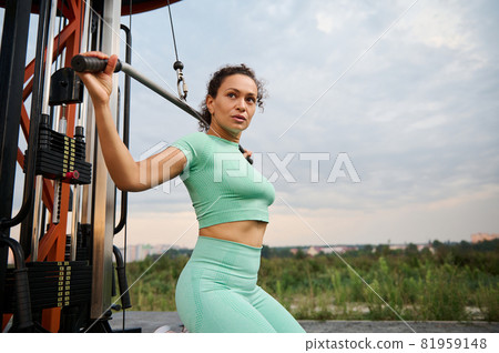 Side view of a Latin American female athlete working out, flexing muscles on a crossover gym equipment in the summer sportsground outdoor in the early morning Side view of a Latin American female athlete working out, flexing muscles on a crossover gym equipment in the summer sportsground outdoor in the early morning 81959148
