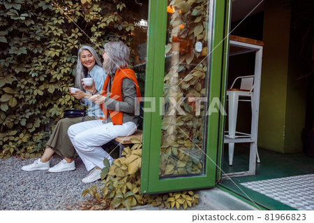 Cheerful senior ladies friends with cups of drinks sit on bench in cafe yard Cheerful senior ladies friends with cups of drinks sit on bench in cafe yard 81966823