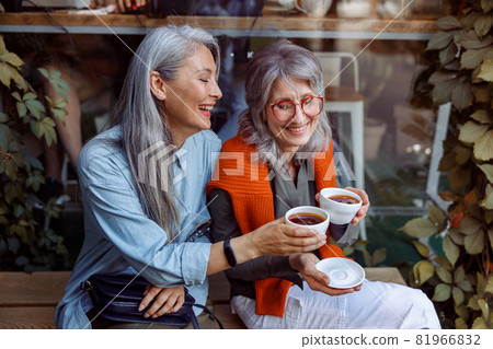 Smiling mature Asian lady with friend clink cups of coffee sitting near cafe Smiling mature Asian lady with friend clink cups of coffee sitting near cafe 81966832