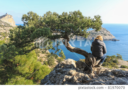 The girl is sitting on the trunk of a juniper tree and looks at the sea. The tree stands on a rock The girl is sitting on the trunk of a juniper tree and looks at the sea. The tree stands on a rock 81970070