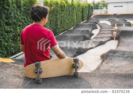Asian man sitting with surfskate or skate board in pumptrack skate Park when sunrise time over photo blur of pumptrack curve, extream sport, healthy and exercise, fashion in covid19 concept 81970236