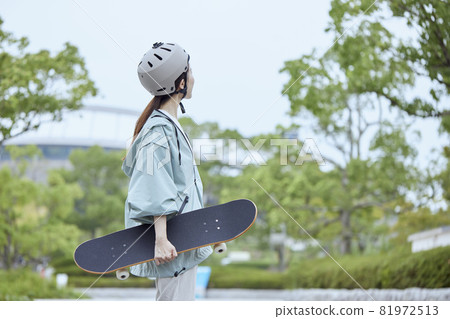 A woman in a park wearing a helmet and holding a skateboard, facing sideways. A woman in a park wearing a helmet and holding a skateboard, facing sideways. 81972513