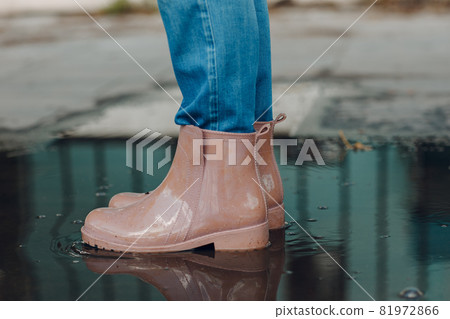 Woman wearing rain rubber boots walking running and jumping into puddle with water splash and drops in autumn rain Woman wearing rain rubber boots walking running and jumping into puddle with water splash and drops in autumn rain 81972866