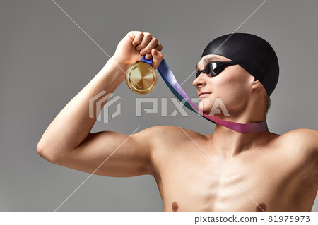 Swimmer with a medal on a gray background rejoices in victory, an athlete in excellent physical shape celebrates a victory clutching a medal in his hand, victory concept, gray background, copy space 81975973
