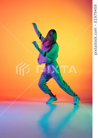 One Caucasian sportive girl dancing hip-hop in stylish clothes on colorful background at dance hall in neon light. Youth culture, movement, style and fashion, action. One Caucasian sportive girl dancing hip-hop in stylish clothes on colorful background at dance hall in neon light. Youth culture, movement, style and fashion, action. 81979409