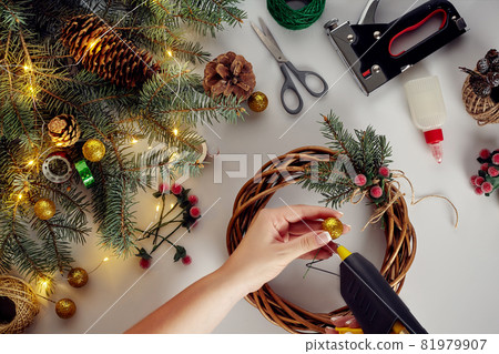 Female hands decorate the Christmas wreath with spruce branches with a red berry and forest cones. White background. New Year's concept. Female hands decorate the Christmas wreath with spruce branches with a red berry and forest cones. White background. New Year's concept. 81979907