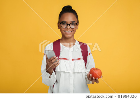Happy smart young afro american girl pupil in glasses with backpack hold smartphone and red apple Happy smart young afro american girl pupil in glasses with backpack hold smartphone and red apple 81984320