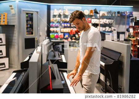 Side view of young male customer in casual clothes playing electronic piano while checking quality of musical instrument during shopping in modern store Side view of young male customer in casual clothes playing electronic piano while checking quality of musical instrument during shopping in modern store 81986282