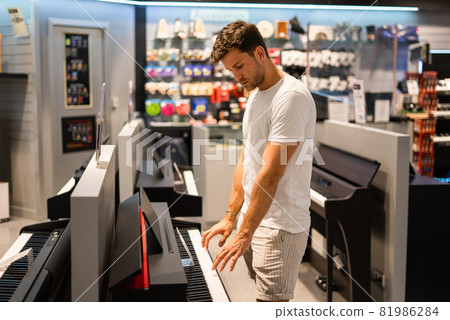 Side view of young male client playing electric piano while visiting modern shop and choosing musical instrument Side view of young male client playing electric piano while visiting modern shop and choosing musical instrument 81986284
