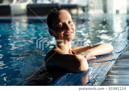 Portrait of young adult caucasian beautiful female model in sport black swimsuit in clear blue water edge indoor swimming pool at dark evening time. Relax harmony and wellness spa therapy Portrait of young adult caucasian beautiful female model in sport black swimsuit in clear blue water edge indoor swimming pool at dark evening time. Relax harmony and wellness spa therapy 81992124