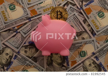 Cute pink plastic piggy bank with bitcoin coin inside on the background of US dollars banknotes. Beautiful photo of a piggy bank and cryptocurrency money. Concept of storing money in digital currency 81992545