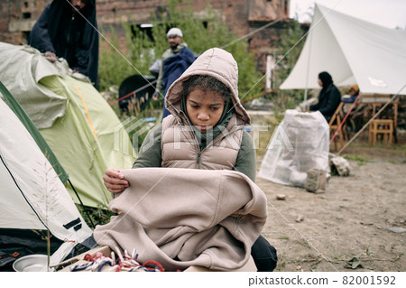 Sad middle-eastern refugee girl in hooded vest holding plaid while freezing in tent camp for migrants 82001592