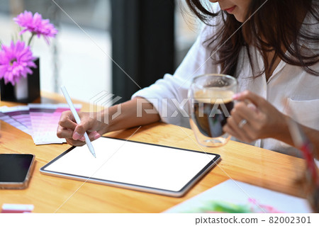 Cropped shot young woman holding coffee cup and working with digital tablet in creative office. Cropped shot young woman holding coffee cup and working with digital tablet in creative office. 82002301