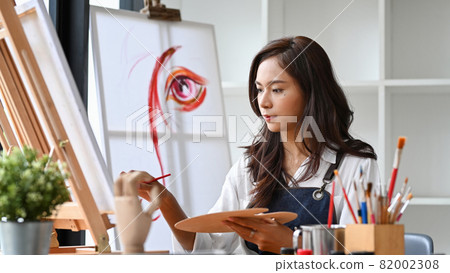 Pretty female artist painting on canvas in bright daylight studio. Pretty female artist painting on canvas in bright daylight studio. 82002308