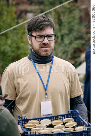 Empathetic male social worker with Volunteer badge carrying box of tin cans for refugees Empathetic male social worker with Volunteer badge carrying box of tin cans for refugees 82003066
