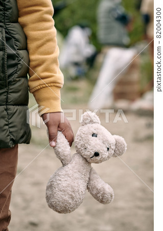 Close-up of unrecognizable refugee child in sweater and vest walking with plush bear through ruins Close-up of unrecognizable refugee child in sweater and vest walking with plush bear through ruins 82004300